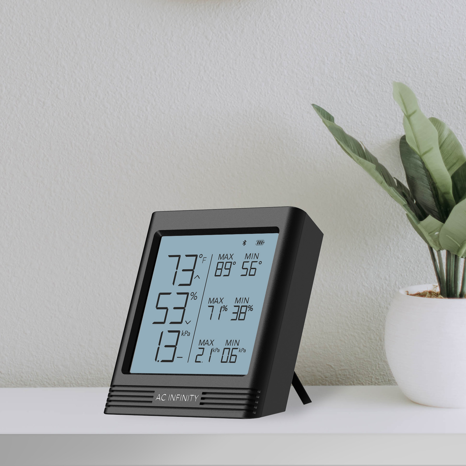 AC Infinity Smart Hygrometer B2 mit beleuchtetem LCD-Display und ausklappbarem Standfuß auf einem Tisch neben einer Zimmerpflanze